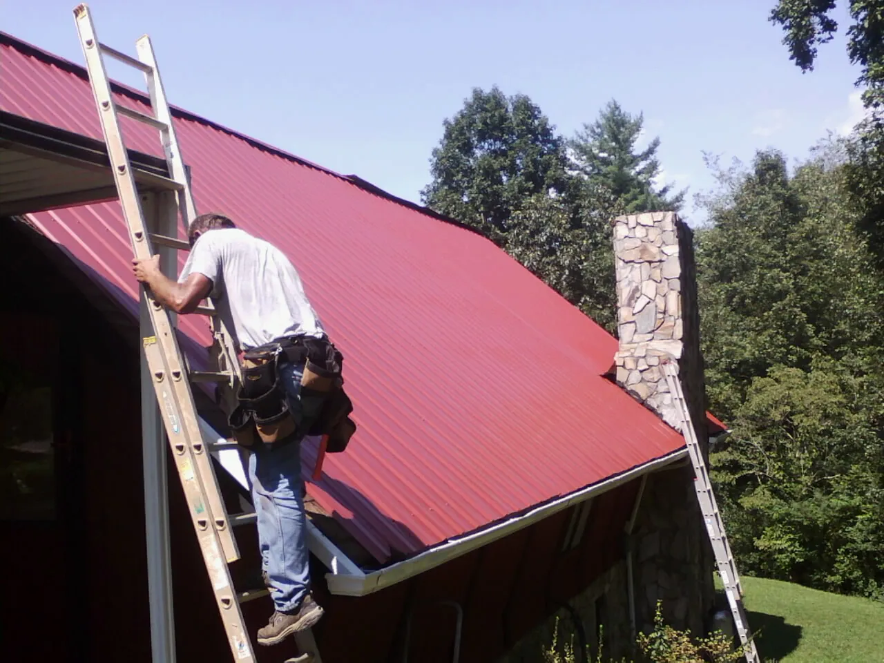 Roofer on a ladder beside a red metal roof and stone chimney — Soffit & Fascia Repair by Roofing Brothers in Reddick
